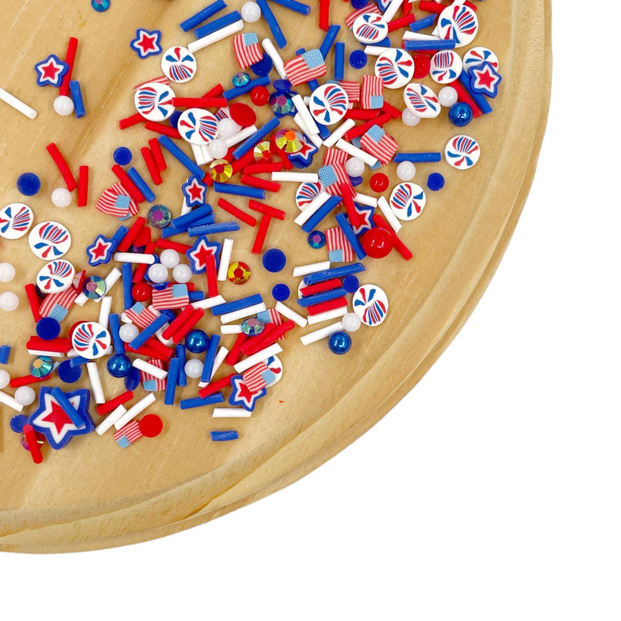 A variety of red, white, and blue polymer clay pieces arranged on a wooden surface, including stars, stripes, and round shapes.

This loose clay mix is sold in a resealable 1 oz Bag

- Material: Polymer Clay and Glitter