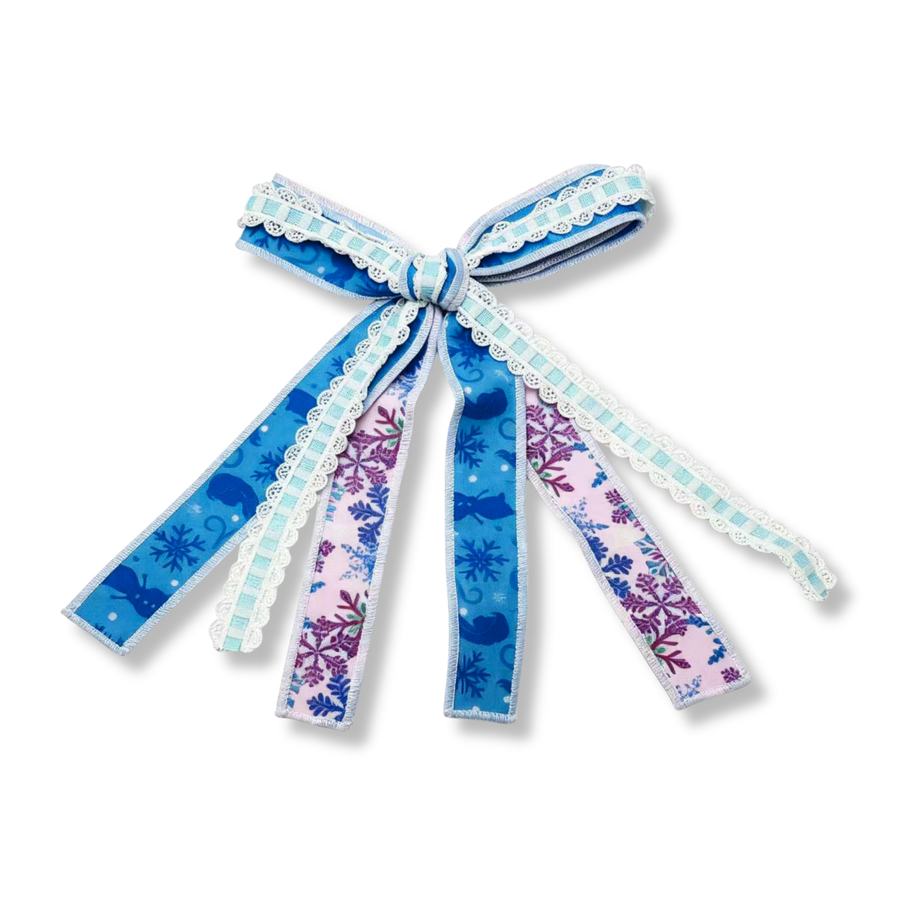 A whimsical ribbon tie bow crafted from three coordinating ribbons featuring winter-inspired blue and lavender prints. The blue ribbons are adorned with snowflakes, swirling accents, and whimsical silhouette details, while the lavender ribbon showcases intricate snowflake patterns in shades of purple and blue. Delicate lace-trimmed accents and flowing tails add beautiful texture and movement, creating a charming storybook-inspired winter design.
Bow Sizing 4" x 8"