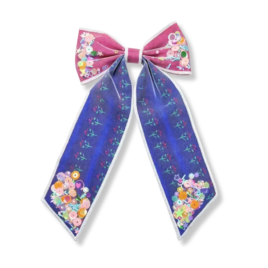 A whimsical long tail shaker bow featuring rich blue and berry pink panels with delicate floral accents and clear shaker pockets filled with colorful sequins, pearls, stars, and confetti. 
Bow Sizing 5" x 8.5"
