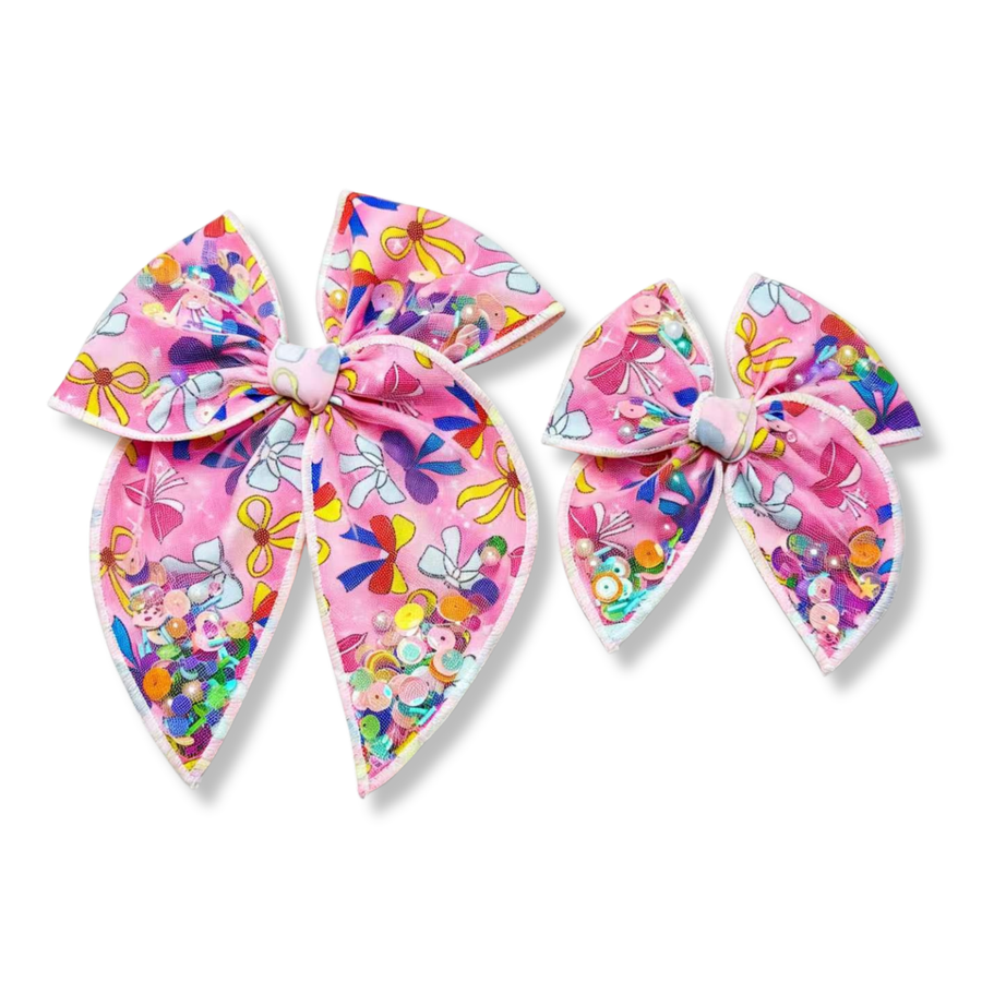 Shaker bow made from pink fabric printed with colorful ribbon and bow designs in shades of yellow, blue, purple, red, and white. The bow features white edging and shaker pockets filled with iridescent sequins, pearls, beads, and confetti in bright pastel tones, creating a playful, vibrant, and sparkly look.
available in two sizes
Small 4.5" x 4.5"
Large 6.5" x 6.5"