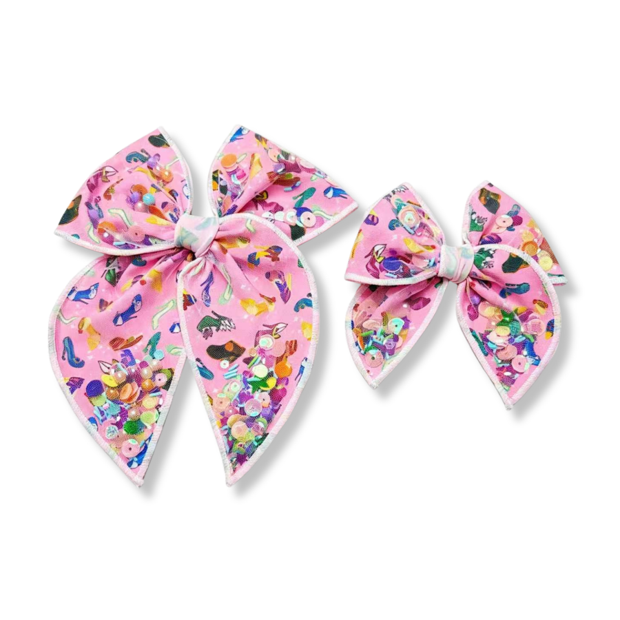 Shaker bow made from pink fabric printed with colorful shoe and accessory designs in shades of blue, yellow, green, purple, red, and black. The bow features white edging and shaker pockets filled with iridescent sequins, pearls, beads, and confetti in bright pastel tones, creating a playful, vibrant, and sparkly look.
Available in two sizes
Small 4.5" x 4.5"
Large 6.5" x 6.5"
