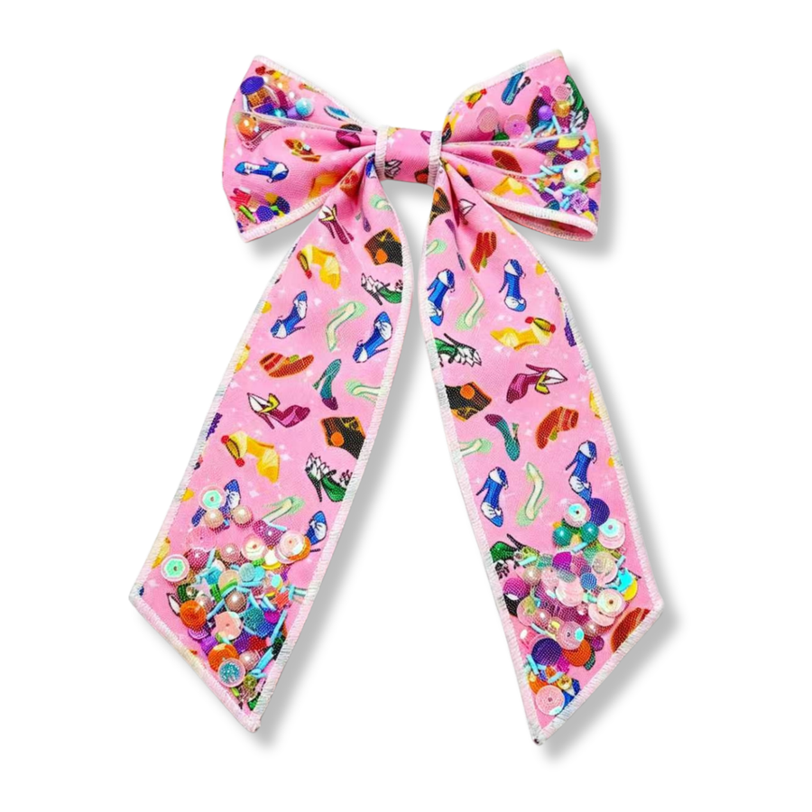 Long tail shaker bow made from soft pink fabric printed with colorful shoe and accessory designs in shades of blue, yellow, green, purple, red, and black. The bow features white edging, long flowing tails, and shaker pockets filled with iridescent sequins, pearls, beads, and confetti in bright pastel tones, creating a playful, vibrant, and sparkly look.
Bow Sizing 5" x 8.5"