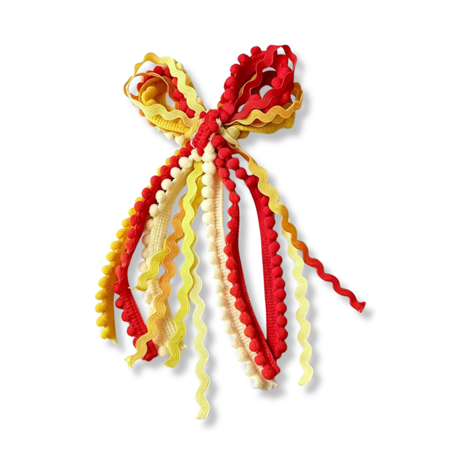 A playful **messy bow** crafted from an assortment of ric rac ribbons, pom-pom trim, and textured woven ribbons in vibrant shades of red, golden yellow, cream, and soft yellow. The cascading ribbons feature wavy edges, plush pom-pom accents, and varied textures that create beautiful movement, dimension, and a whimsical handcrafted look. The warm, cheerful color palette gives this bow a bright and playful charm, making it a fun statement accessory full of personality. 
Bow Sizing 5" x 9"