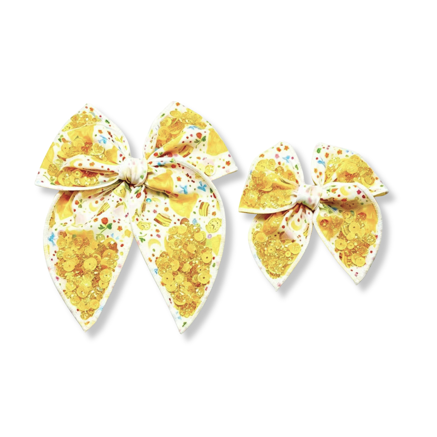 This whimsical princess-inspired bow features a white background decorated with yellow ball gowns, enchanted clocks, slices of cake, bows, flowers, and other fairytale-themed details. Clusters of shimmering gold sequins are incorporated throughout the bow, creating eye-catching sparkle across both the loops and tails. The bow is available in both a larger and smaller size, each featuring softly rounded loops and pointed tails for a classic, elegant silhouette.
Small 4.5" x 4.5"
Large 6.5" x 6.5"

