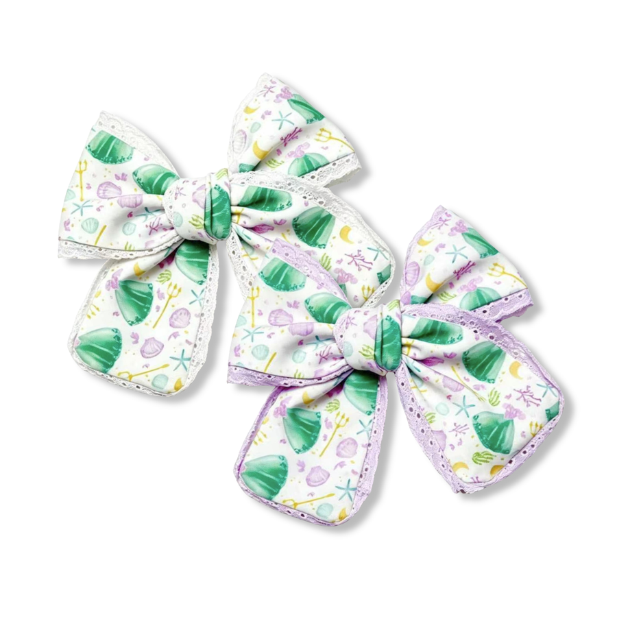 These  boutique bows feature a whimsical under-the-sea print with seafoam green mermaid inspired dresses, lavender seashells, starfish, bubbles, and ocean-inspired details on a soft white background. Delicate white or lavender lace trim accents the edges, adding elegant texture and dimension to the design. The bow silhouette and dreamy pastel color palette create a magical mermaid-inspired accessory full of seaside charm and storybook beauty. 
You choose white or lavender lace trim
SIZE:
Small 5" X 6"


