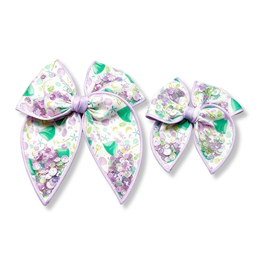 These enchanting shaker bows feature a whimsical under-the-sea print with seafoam green mermaid inspired dresses, lavender seashells, starfish, bubbles, and ocean-inspired details on a soft white background. Filled with a sparkling mix of pastel lavender, mint, and iridescent sequins, the shaker accents add beautiful movement and shimmer. 

you choose size
Small 4.5" x 4.5"
Large 6.5" x 6.5"