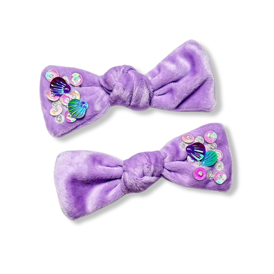 These adorable bows are crafted from soft lavender plush fabric with a cozy, velvety texture. Each bow is accented on one side with iridescent sequins, sparkling gems, and colorful shell embellishments that add a whimsical under-the-sea touch. The combination of the plush material and shimmering mermaid-inspired details creates a playful, magical accessory full of charm and sparkle. 
MEASUREMENTS

2" x 5"

Set of 2

