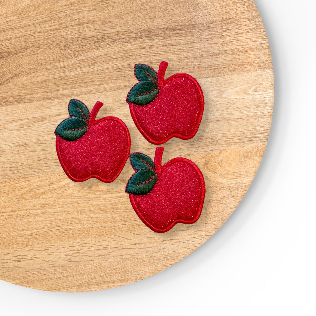 Embroidered red apple iron-on patch featuring a bright red apple with a subtle glittery finish, accented by two dark green leaves and a short stem. Finished with smooth satin stitching and bold edging for a vibrant, classic fruit design. 
APPROX 1.5" Tall X 2" Wide&nbsp;