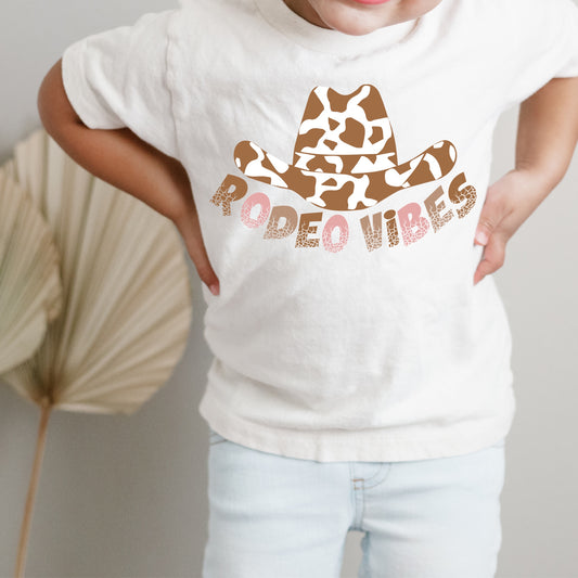 Brown and white cow print cowgirl hat and the phrase "Rodeo Vibes" in pink and brown lettering.