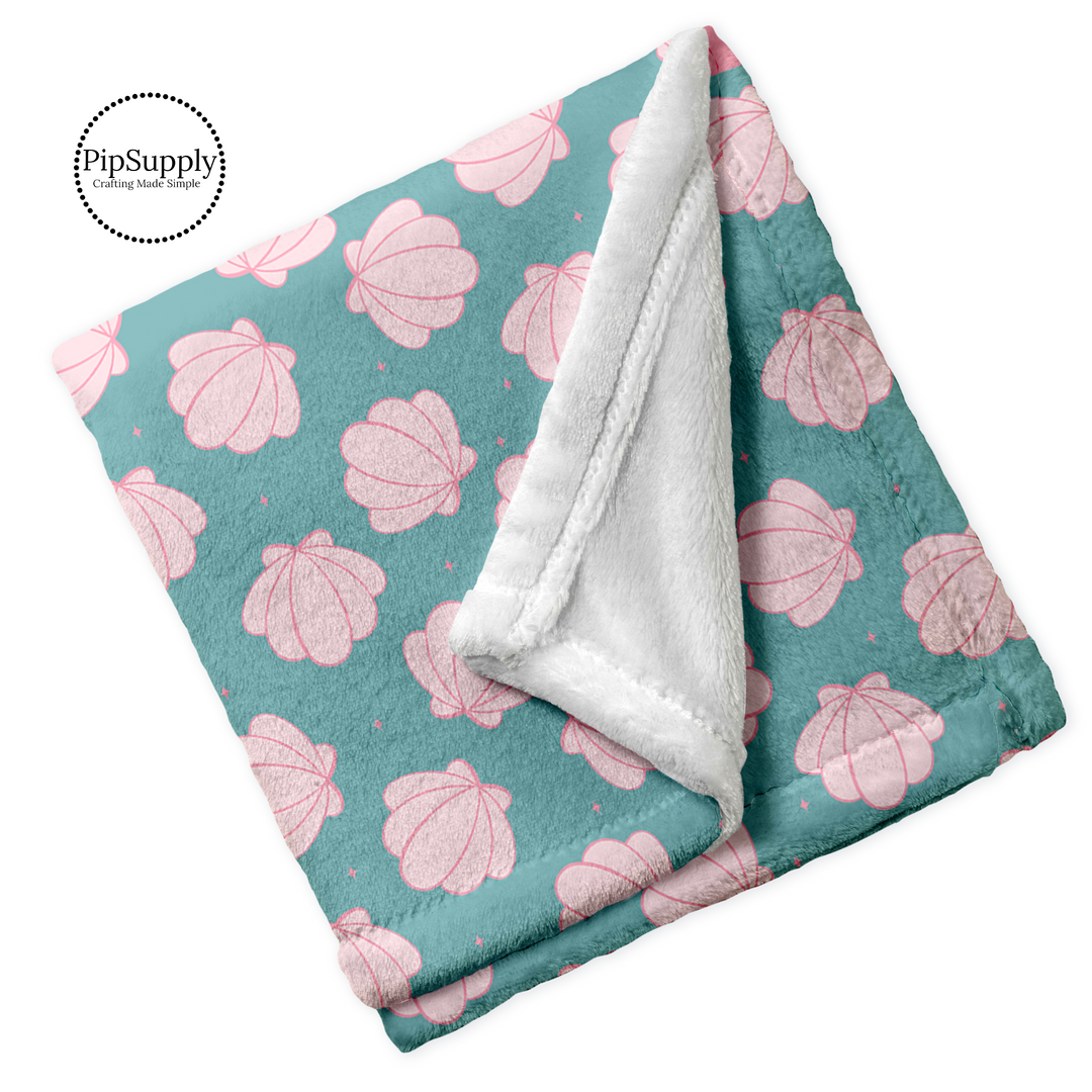 Soft plush white folded blanket with top printed with light pink sea shells clams on aqua background.