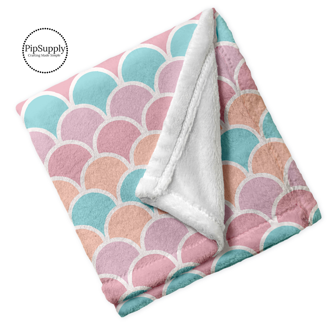 Soft folded white blanket with pastel coral, orange, aqua, and pink mermaid scale pattern.