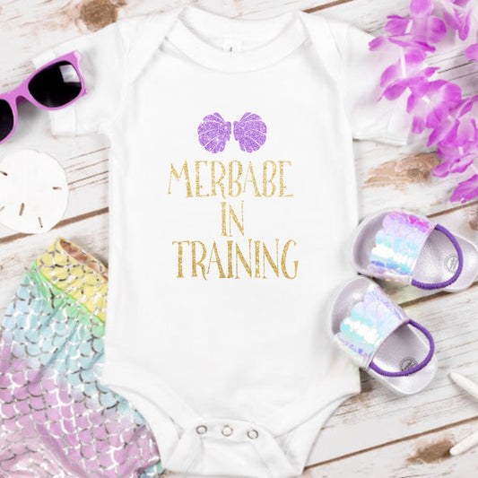 "Merbabe in training" in gold glitter font and purple sparkly seashells
