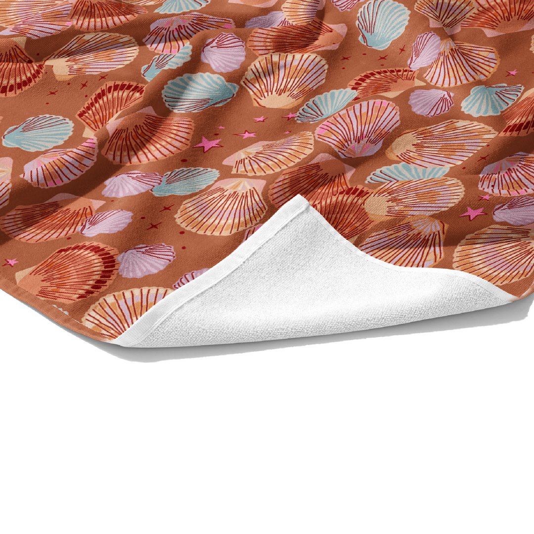 Plush white cotton towel with top printed with brown outlined seashells in light blue, lavender, and tan on a brown background.